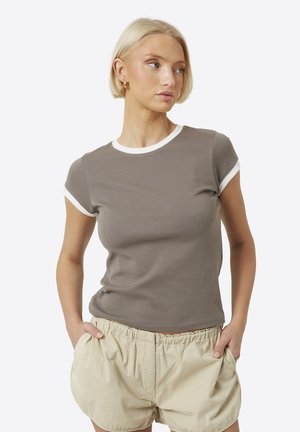 Vrouw met kort blond haar, gekleed in een taupekleurig t-shirt met witte bies en beige korte broek, staand met handen in de zakken, kijkend naar rechts.