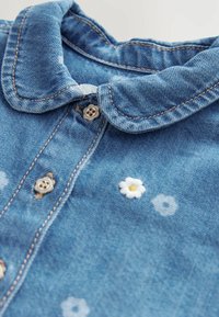 Chemise en denim avec des boutons en forme de fleurs et de petites marguerites blanches brodées, présentant un col arrondi et des coutures visibles.