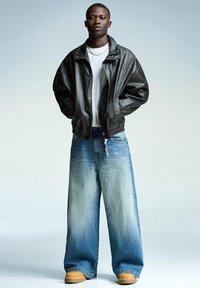 Jachetă bomber din piele neagră, tricou alb, blugi supradimensionați de culoare albastru deschis, pantofi cu platformă maro; modelul stă cu mâinile în buzunare pe un fundal gradient.