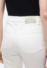 Κοντινό πλάνο ενός ατόμου που φοράει λευκά τζιν παντελόνια με πίσσω τσέπες και ετικέτα Calvin Klein, σε συνδυασμό με μαύρο τοπ.