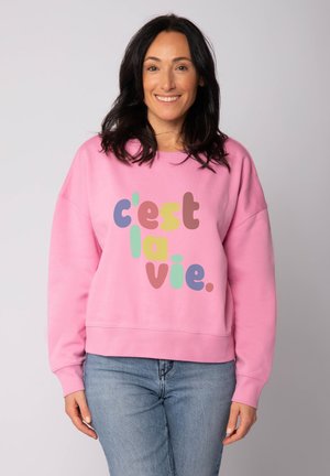 Rosa Sweatshirt mit Rundhalsausschnitt, versehen mit mehrfarbigem Text "c'est la vie." Weiche Textur und lockere Passform, kombiniert mit hellblauen Jeans.