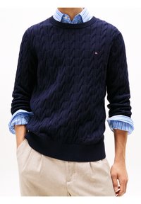 Pull en maille torsadée bleu marine avec un col et un bas côtelés, superposé sur une chemise rayée bleu clair. Texture douce ; coupe décontractée.