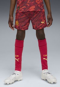 Maglia a maniche corte con motivo rosso e arancione, abbinata a pantaloncini rossi con un logo. Include calze rosse con dettagli dorati e scarpe bianche.