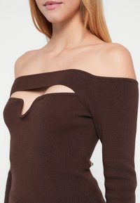 Καφέ τοπ με ακάλυπτους ώμους από ριμπ ύφασμα, με σχέδιο κόψιμο σε σχήμα κλειδιού στο στήθος και εφαρμοστά μακριά μανίκια για ένα σιλουέτα που αγκαλιάζει το σώμα.