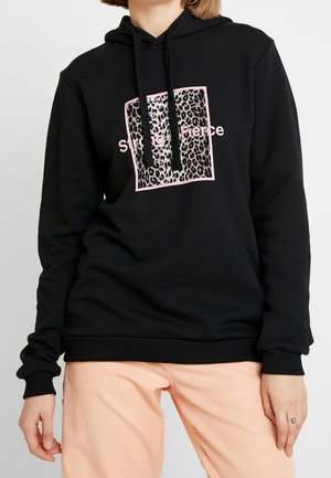 Czarna bluza z kapturem z kwadratem w panterkę z różową obwódką i napisem „Strong Fierce”, w połączeniu z jasnoróżowymi spodniami na stojącej osobie.