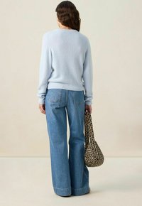 Femme aux cheveux bruns ondulés portant un pull bleu clair et un jean large bleu, tenant un sac à main en imprimé léopard, de dos.