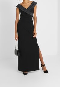 Černé večerní šaty s výstřihem do V, krátkými rukávy a nařasením. Obsahují rozparek po stehna, nošeny s černými podpatky a malou kabelkou přes rameno.