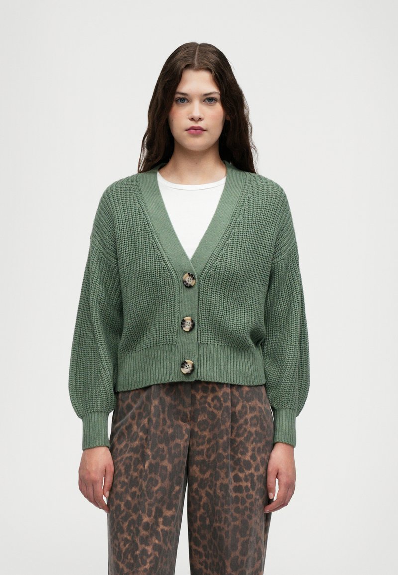 Grønn strikket cardigan med V-hals, tre store knapper og vide ermer, kombinert med brune bukser med leopardmønster.