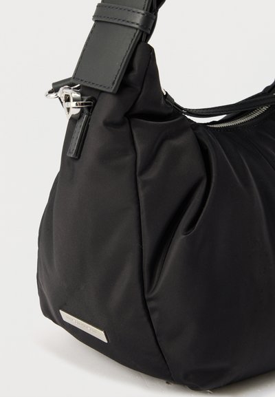 Μαύρη νάιλον τσάντα ώμου με ασημένιο φερμουάρ, ρυθμιζόμενο ιμάντα, πλαϊνή τσέπη και μικρή ορθογώνια μεταλλική ετικέτα με το λογότυπο.