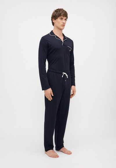 Navy zils pidžamu komplekts, kas sastāv no garām piedurknēm augšdaļas ar kabatu un kontrastējošu baltu apmali, saskaņots ar elastīgām biksēm ar auklu.