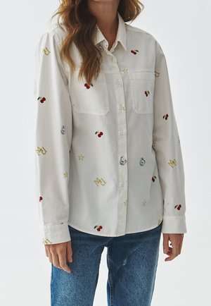 Chemise blanche boutonnée ornée de broderies colorées de cerises, d'étoiles et de couronnes. Manches longues avec poignets et deux poches poitrine.