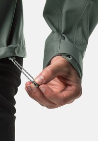 Giacca tecnica verde con finitura testurizzata; mano che tira il laccio dall'orlo, mostrando un cordino grigio e nero con un fermaglio.