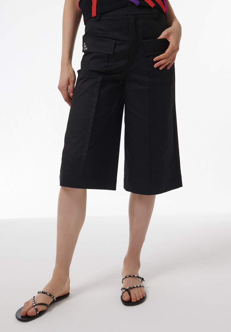 Pantalones culotte negros de tela suave, con grandes bolsillos delanteros y un diseño recortado. Combinados con sandalias negras adornadas con acentos blancos.