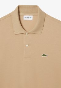 Beige pikétröja i texturerad tyg, med klassisk krage, två knappar och en liten grön krokodil-logotyp på vänstra bröstet.