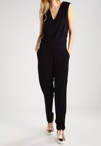 Svart jumpsuit med djup V-ringning, utan ärmar, elastiskt midjeband och sidofickor, kombinerad med roséguld sneakers. Slät tygtextur.