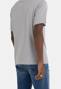 Hombre visto desde atrás con una camiseta de manga corta gris clara y jeans de mezclilla azules, de pie con el brazo izquierdo relajado a su lado.