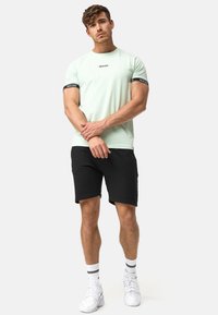 Camiseta de manga corta verde claro con un logo negro, combinada con pantalones cortos de chándal negros. Zapatillas blancas con detalles en negro y calcetines a rayas.