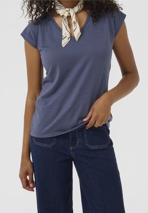 Femme portant un haut bleu à col en V manches courtes, un jean bleu foncé et un foulard blanc à motifs autour du cou, avec des cheveux bouclés visibles.