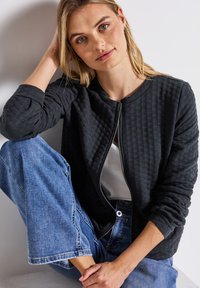 Ung kvinna med blont hår som sitter med ett knä uppdraget, klädd i en texturerad svart zip-up jacka, vit topp och blå denim jeans.