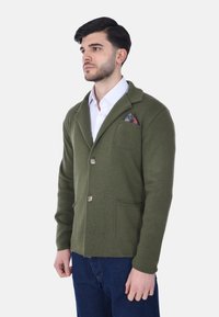 Cardigan verde oliva testurizzato con colletto a scialle, due tasche frontali e tre bottoni. Presenta un fazzoletto da taschino a motivo.