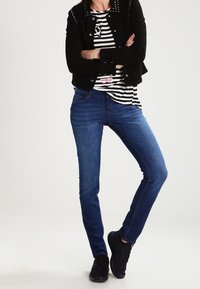 Tmavě modré džíny s úzkým střihem, doplněné černou surovou bundou s kovovými cvoky a pruhovanou košili pod ní. Černé tenisky.