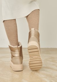 Botas de tobillo beige con un acabado suave, forro acolchado y suelas gruesas. El diseño presenta una textura acolchada y un pequeño lazo trasero.