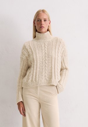Crème kleurige gebreide coltrui met een kabelpatroon. Dikke textuur, losse pasvorm, cropped lengte, gecombineerd met ribbelige beige broeken.