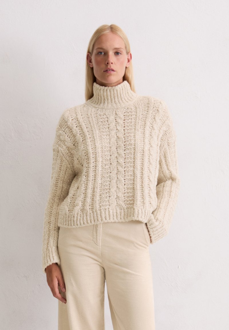 Crème kleurige gebreide coltrui met een kabelpatroon. Dikke textuur, losse pasvorm, cropped lengte, gecombineerd met ribbelige beige broeken.
