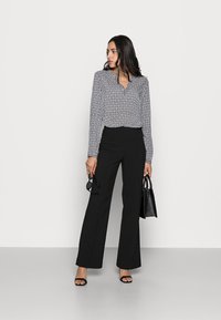 Blusa a maniche lunghe a fantasia bianco e nero con colletto, abbinata a pantaloni neri a vita alta. Gli accessori includono occhiali da sole e una borsa nera.