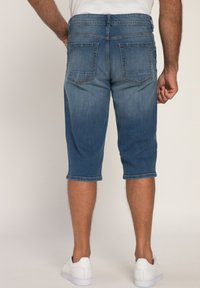 Denim bermuda shorts, kesan biru medium, desain memudar, panjang hingga lutut, lima kantong, detail jahitan di bagian hem, dipadukan dengan sneakers putih.