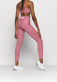 Leggings atléticos rosas y sujetador deportivo a juego con tirantes entrelazados. Presenta el logo de PUMA en la cintura. Usados con zapatillas blancas.