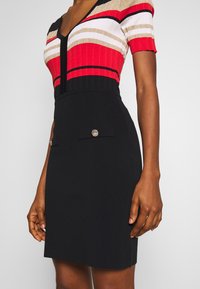 Femme portant une robe à manches courtes avec un col en V, des rayures rouges, blanches et dorées sur le haut et une jupe noire unie avec deux poches avant.