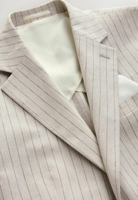 Beige Streifenblazer mit Revers und weißem Taschentuch auf weichem Stoffhintergrund.