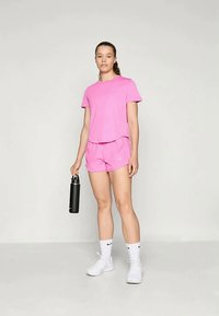 Mulher em camiseta atlética rosa e calções, segurando uma garrafa de água preta, vestindo meias brancas da Nike e sapatilhas brancas, de pé contra um fundo simples.
