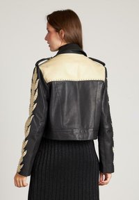 Vrouw van achteren gezien, gekleed in een zwart-beige leren jasje met studs en vlamachtige patronen op de mouwen, en een zwarte rok.