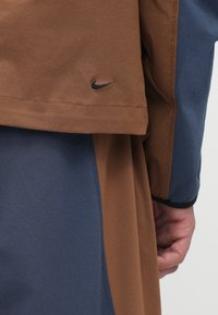 Bruine en marineblauw kleurgeblokkeerde kleding met een gladde textuur; beschikt over een zwart Nike-logo. Inclusief een contrasterend ontwerp voor mouwen en lichaam.