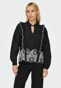 Blouse noire avec un col à volants, une ouverture en forme de clé et des détails en dentelle florale blanche à l'ourlet. Manches longues avec des poignets ajustés.