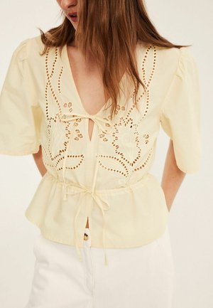 Hellgelbe Bluse mit Puffärmeln, floraler Cutout-Stickerei und vorderen Bindebändern am Ausschnitt und in der Taille, getragen zu weißen Hosen.