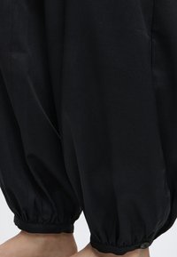 Personne sans jambes portant un pantalon noir ample avec des poignets élastiques rassemblés aux chevilles, debout pieds nus sur un fond blanc.