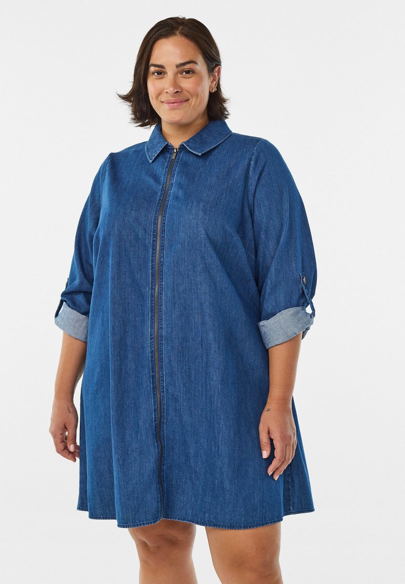 En kvinna ler, klädd i en denimklänning med krage och uppvikta ärmar, mot en enkel vit bakgrund.