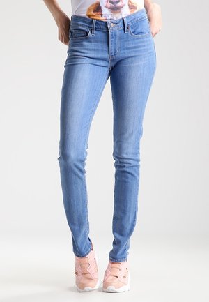 Kvinde iført lyseblå tætsiddende jeans, hvid t-shirt med hundemotiv og pinke sneakers, stående mod en ensfarvet baggrund.