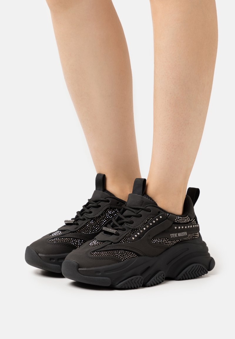 Personne portant des baskets chunky noires Steve Madden avec des détails cloutés et des semelles épaisses texturées sur fond blanc.