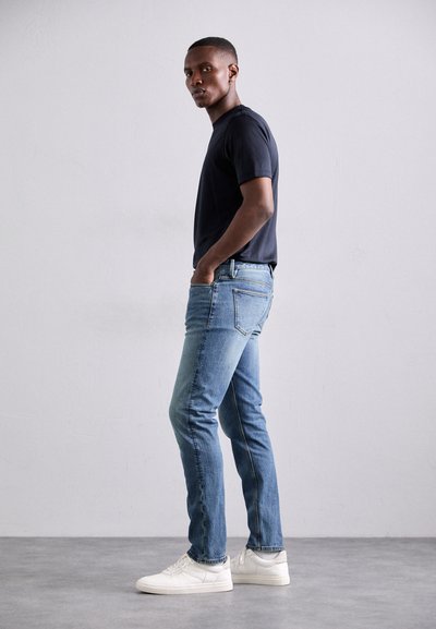 Fekete, rövid ujjú póló, világoskék, slim-fit farmernadrággal és fehér sportcipőkkel, semleges szürke háttér előtt állva.