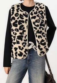 Person som bär en ärmlös beige och svart leopardmönstrad knapplös väst över en svart långärmad topp och blå jeans med en brun axelrem.