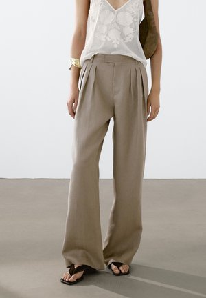 Person trägt beige weite Hosen, weißes ärmelloses Top mit Spitzenmuster, goldenes Armreif und schwarze Sandalen und steht auf grauem Boden.