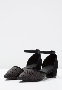 Černé boty s perforovaným koženým svrškem, špičatou špičkou, řemínkem kolem kotníku a nízkým blokovým podpatkem; obsahují hladké a strukturované materiály.