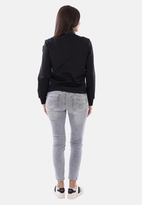 Mujer de pie de espaldas vistiendo una chaqueta negra, jeans pitillo gris claro y zapatillas blancas con detalles negros.