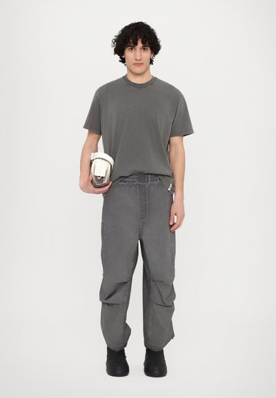 Grijze katoenen T-shirt en losse grijze broek. De outfit heeft een ontspannen ontwerp met zichtbare stiksels. Model houdt een witte tas vast.