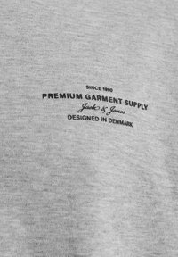 Siva bombažna majica z black vezenim besedilom, ki vključuje "Premium Garment Supply" in "Jack & Jones," z gladko teksturo.