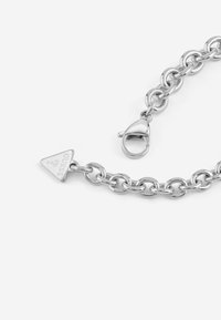 Bracciale in argento con maglie ovali intrecciate, un ciondolo triangolare con la scritta "Guess" e chiusura con gancio a moschettone.